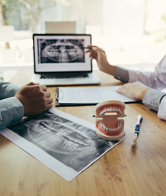 The image shows two individuals seated at a table with a laptop, papers, and a model of a tooth on a stand, discussing dental care with a projected medical image visible behind them.