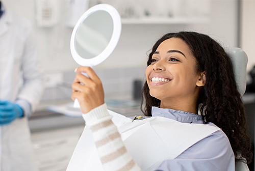 A woman in a dental chair holding up a mirror to her face with a smile.
