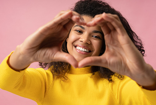 The image shows a person making a heart shape with their hands against a pink background.